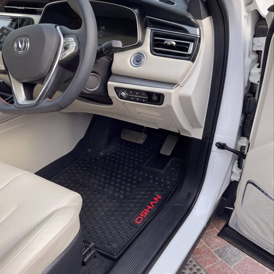 A black rubber car floor mat with red 'OSHANI' branding is shown installed in the driver's footwell of a white vehicle. The interior features beige leather seats and a dark dashboard.