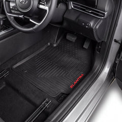 Black rubber car floor mats installed in the front footwells of a Hyundai Elantra, with the red 'ELANTRA' logo printed on the passenger side mat.