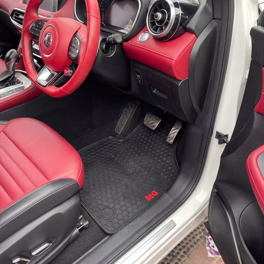 Black rubber car floor mats with a honeycomb pattern and a small red 'MG' logo, shown installed inside a red and black MG car with the door open.