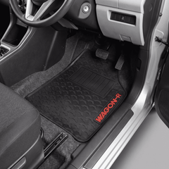 A black car floor mat with a wavy quilted pattern and a red 'WAGON R' logo, shown installed in the driver's seat of a vehicle.