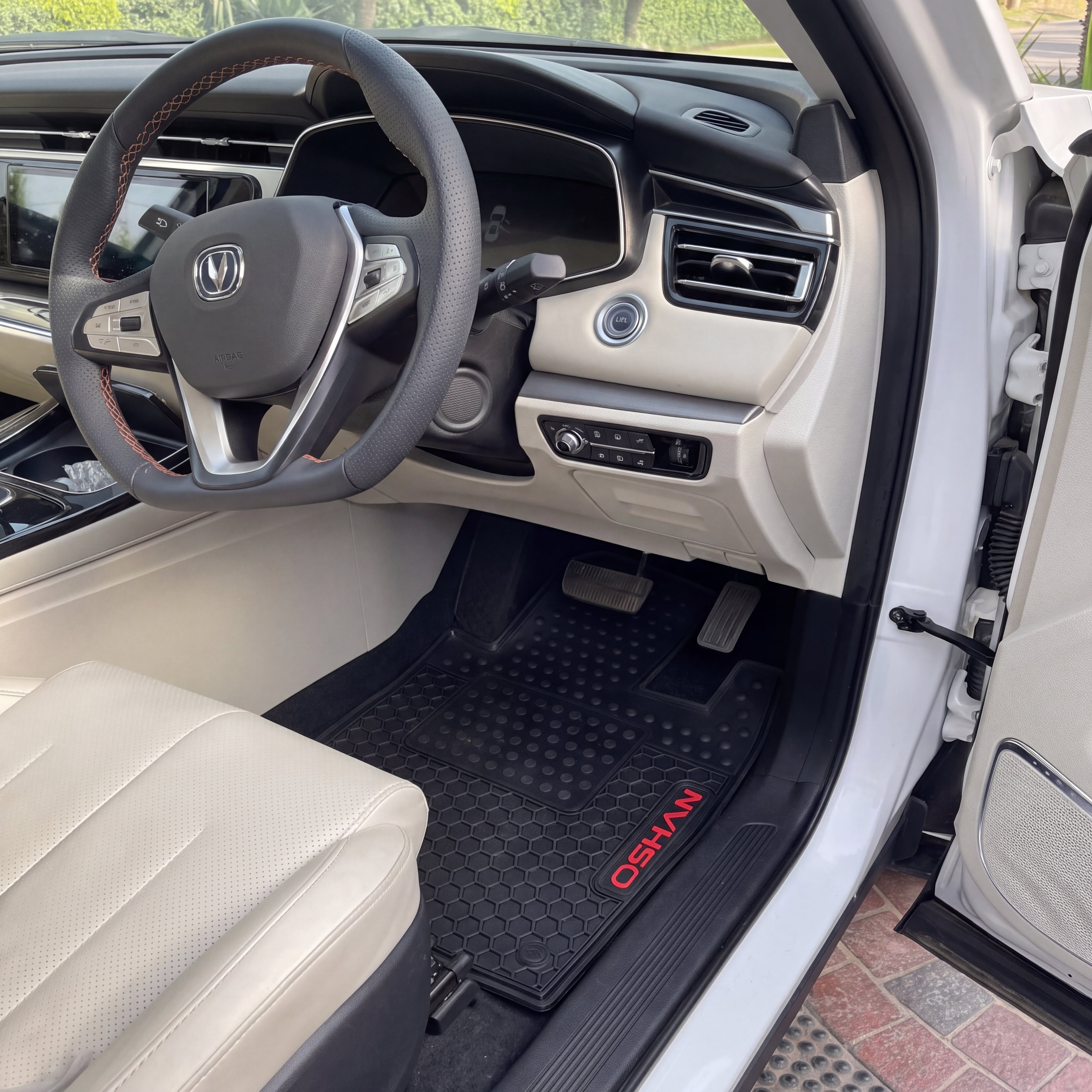 A black rubber car floor mat with red 'OSHANI' branding is shown installed in the driver's footwell of a white vehicle. The interior features beige leather seats and a dark dashboard.