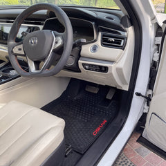 A black rubber car floor mat with red 'OSHANI' branding is shown installed in the driver's footwell of a white vehicle. The interior features beige leather seats and a dark dashboard.