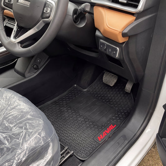 Black rubber car floor mats installed in the front of a vehicle, with the driver's seat visible and a HAVOL logo on the mat. The interior features a gray steering wheel, a tan dashboard panel, and a seat covered in protective plastic.