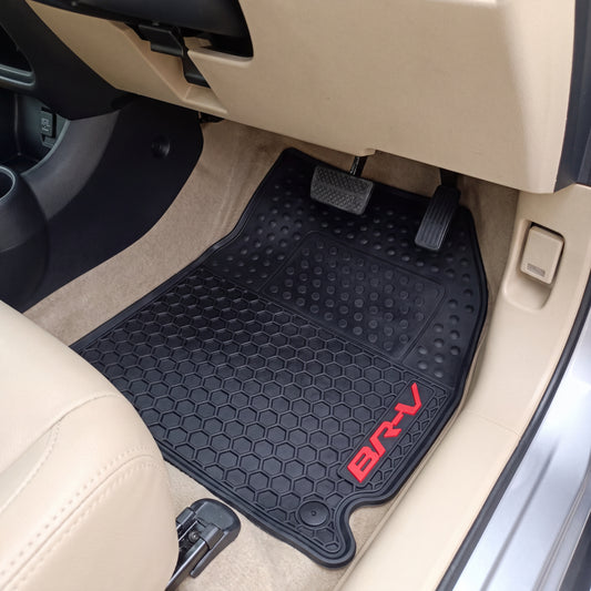 A black textured car floor mat with a red 'BRV' logo, shown installed in the driver's footwell of a vehicle with beige interior trim