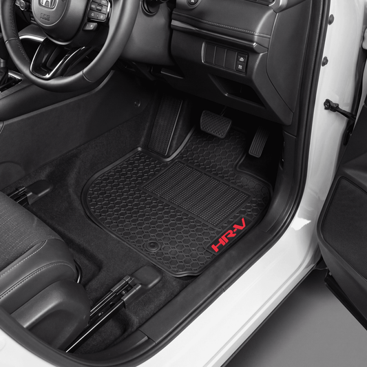 Black textured car floor mats installed in a vehicle's footwell, with a red embroidered 'HRV' logo on the driver's mat and the door open showing the white exterior.