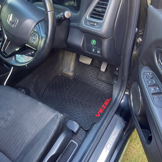 Black car floor mat with red 'VEZEL' text, installed in the driver's footwell of a Honda vehicle with the door open.