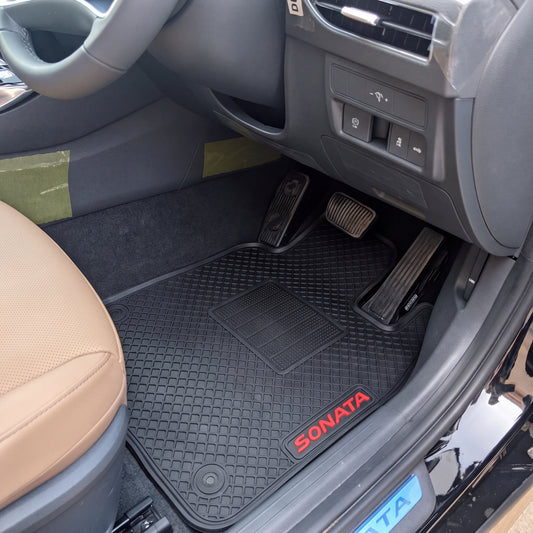 Black rubber car floor mats installed in a vehicle interior, with a red SONATA logo on the front mat and a beige driver's seat visible.