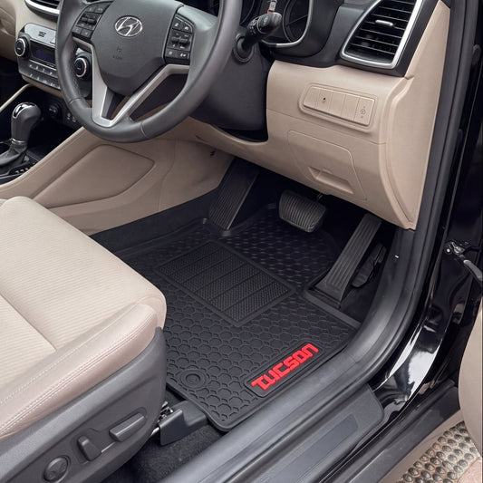 A pair of black Tucson floor mats with red 'TUCSON' branding, shown installed inside a beige Hyundai Tucson vehicle with the driver's side door open.