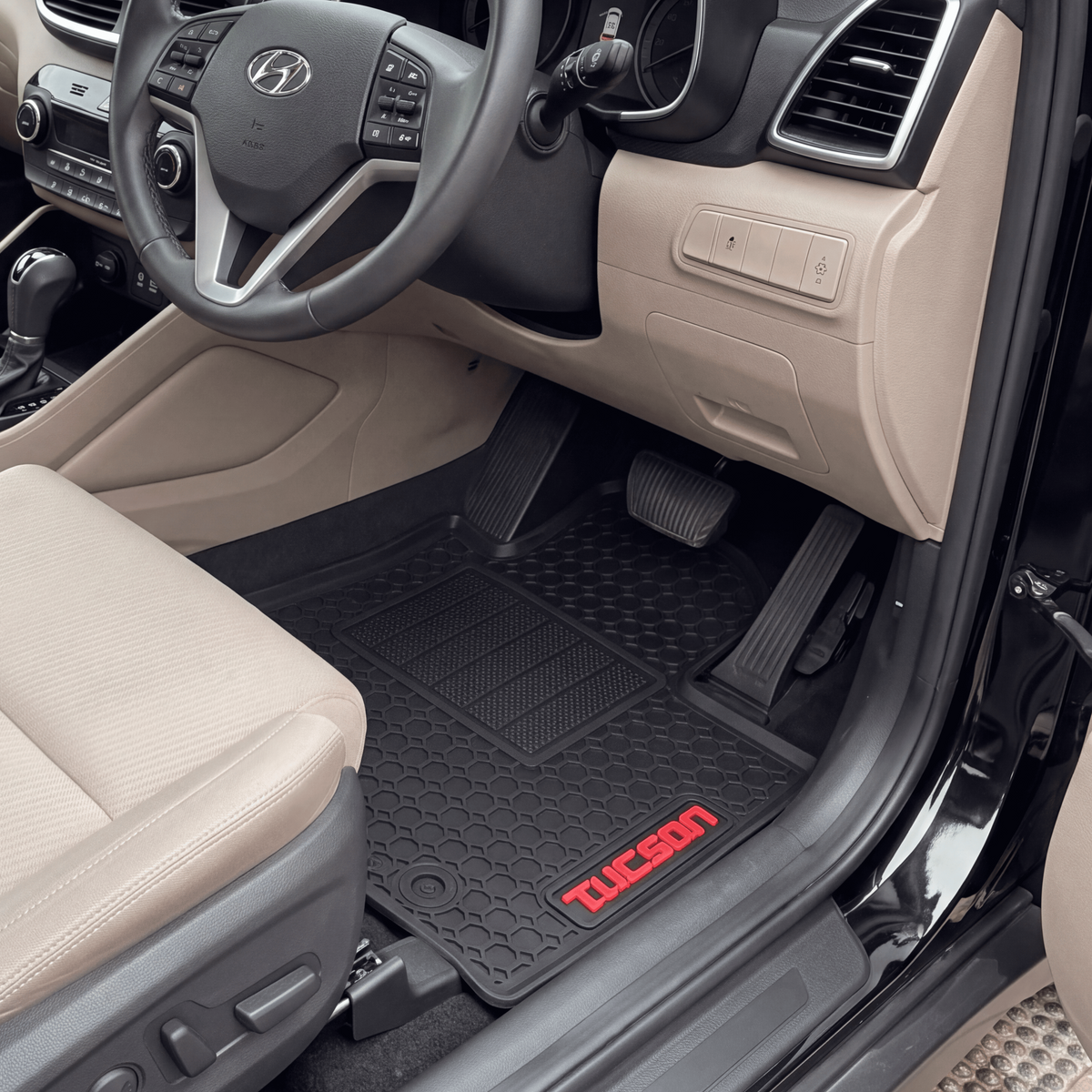 A pair of black Tucson floor mats with red 'TUCSON' branding, shown installed inside a beige Hyundai Tucson vehicle with the driver's side door open.