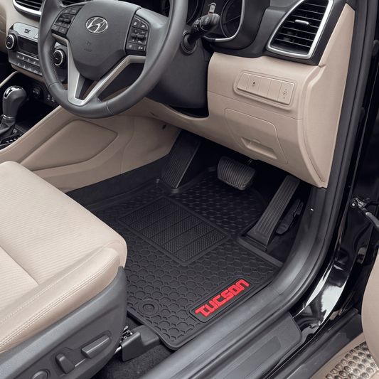 A pair of black Tucson floor mats with red 'TUCSON' branding, shown installed inside a beige Hyundai Tucson vehicle with the driver's side door open.