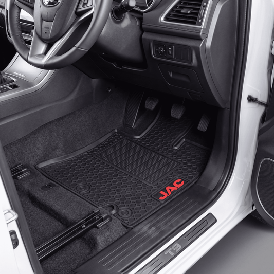 A black rubber car floor mat with a honeycomb pattern and a red JAC logo, shown installed in the driver's seat of a vehicle with the door open.