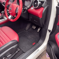 Black rubber car floor mats with a honeycomb pattern and a small red 'MG' logo, shown installed inside a red and black MG car with the door open.