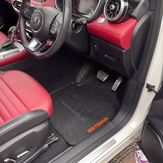 MG HS luxury car interior with a black textured latex floor mat on the driver's side, featuring an orange MG TROPHY logo. The car is shown with red leather seats and black dashboard, with a text overlay at the bottom reading 'MG HS TROPHY LATEX CAR FLOOR MAT'.