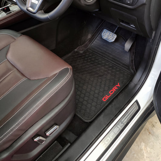 Black car floor mat with a honeycomb pattern and red 'GLORY' branding, shown installed inside a vehicle next to the brown leather seat.