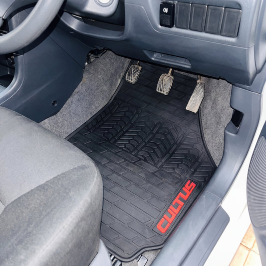 A black rubber car floor mat with a red COTUS logo, shown installed in the driver's footwell of a gray vehicle.