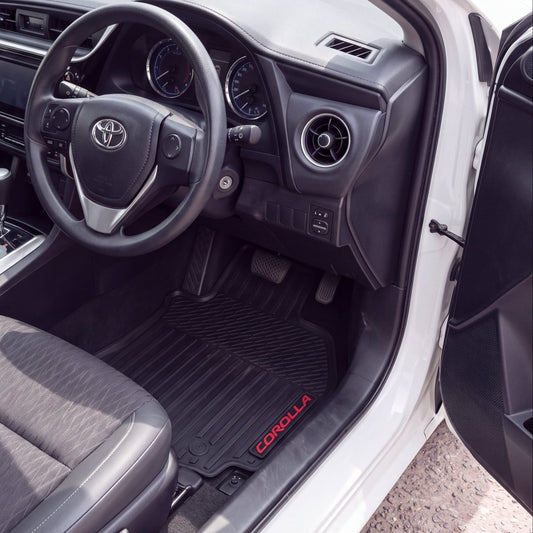 Black Corolla rubber floor mats installed in the front cabin of a white Toyota sedan, shown with the driver's side door open.