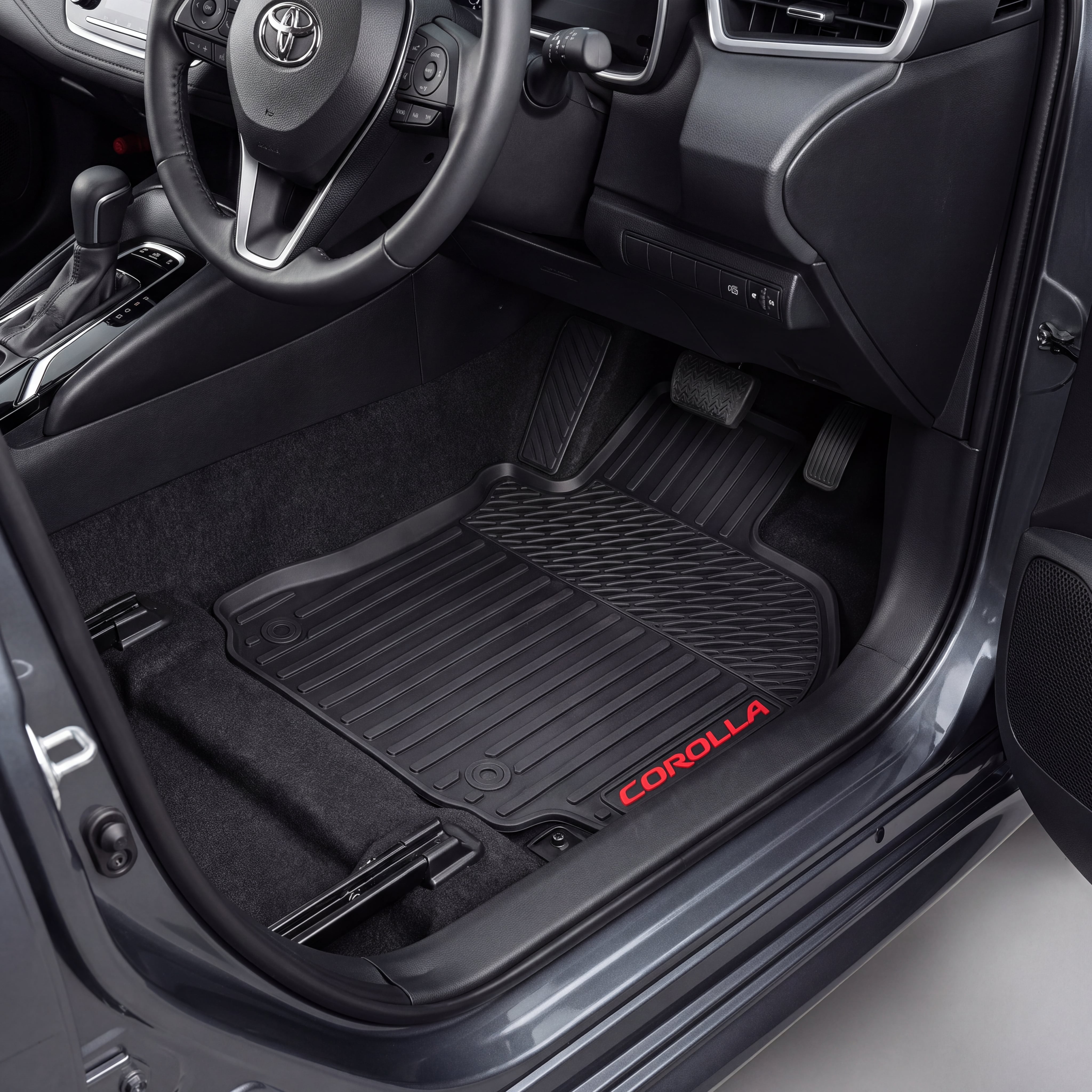 Black car floor mats installed in a Toyota Corolla, shown from the open driver's side door. The mats are textured and black, with the red 'COROLLA' logo embroidered on the lower edge.