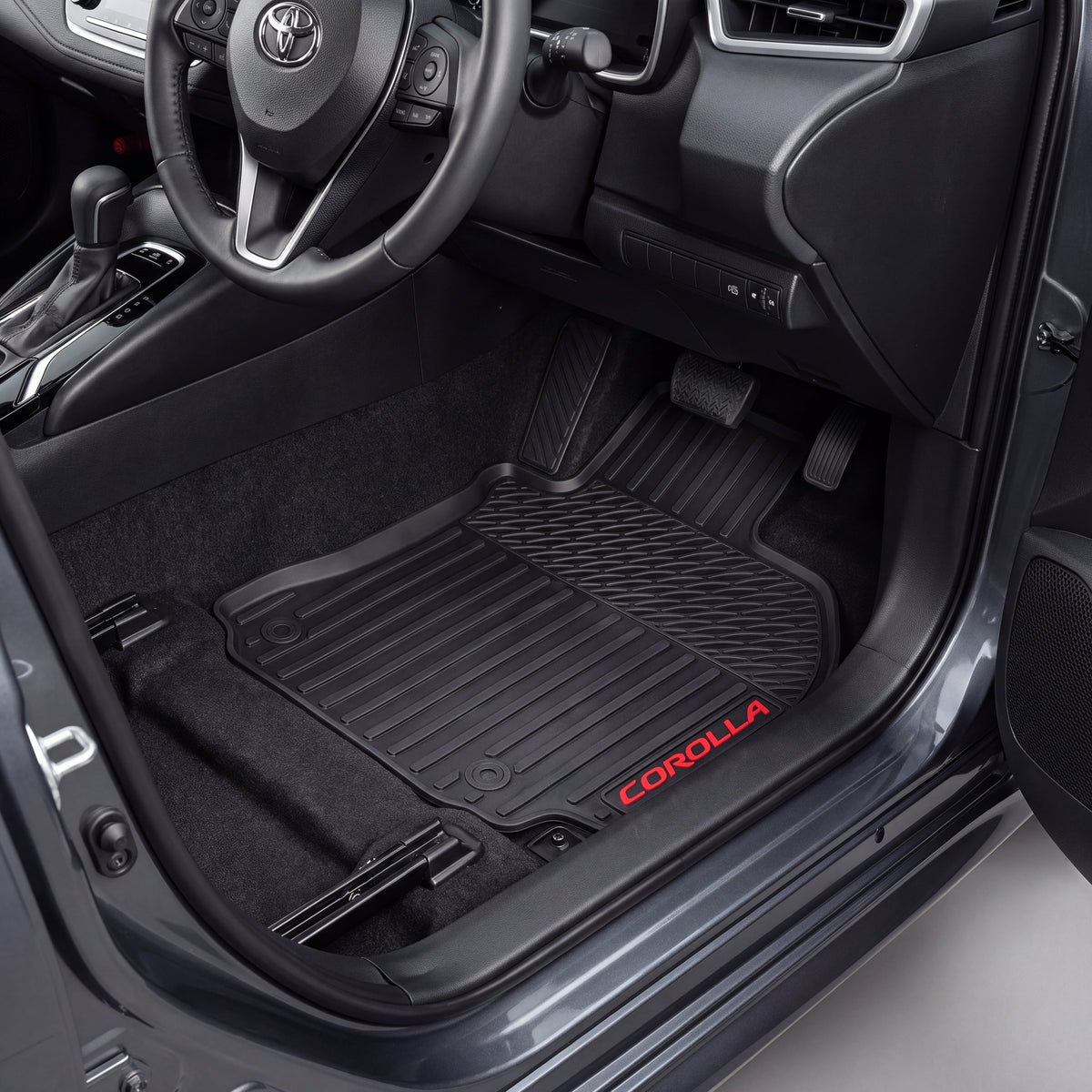 Black car floor mats installed in a Toyota Corolla, shown from the open driver's side door. The mats are textured and black, with the red 'COROLLA' logo embroidered on the lower edge.