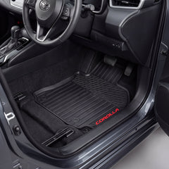 Black car floor mats installed in a Toyota Corolla, shown from the open driver's side door. The mats are textured and black, with the red 'COROLLA' logo embroidered on the lower edge.