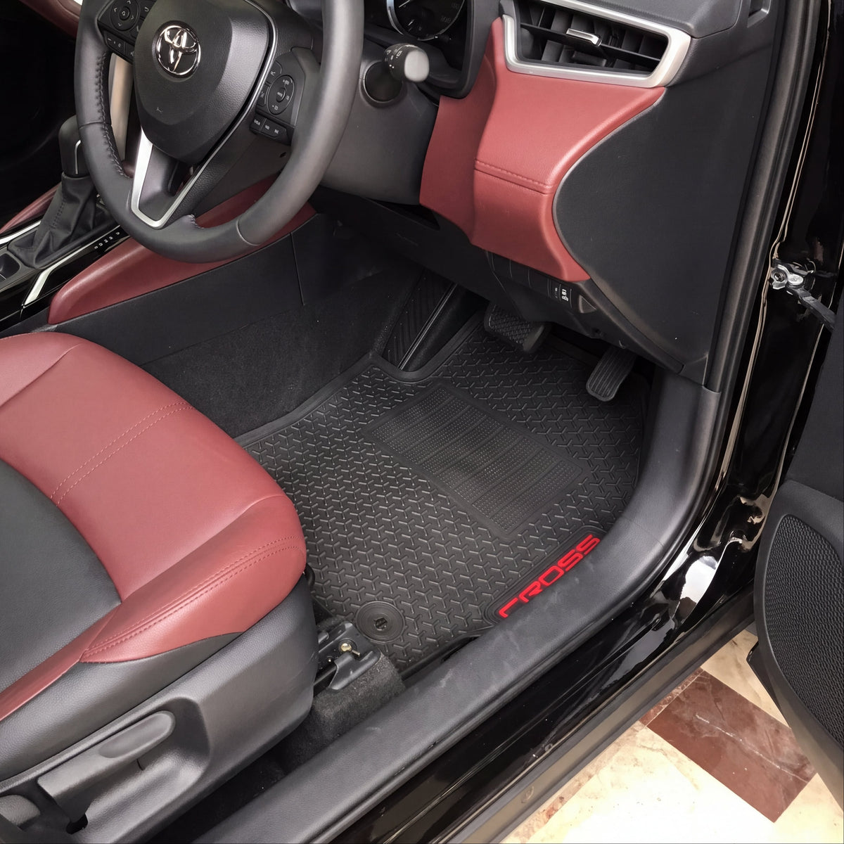 A car interior with black and burgundy leather seats, a black steering wheel with a silver Kia logo, and black textured floor mats with red 'PROSE' branding.