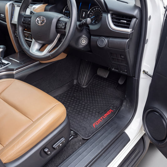 Black rubber car floor mats with a diamond-embossed pattern and red FORTUNER branding, shown installed in the driver's side of a vehicle with a tan leather seat and black interior.