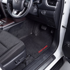 Black rubber car floor mats with a diamond-patterned texture and red 'FORTUNER' branding, shown installed in the driver's seat of a vehicle with the door open.