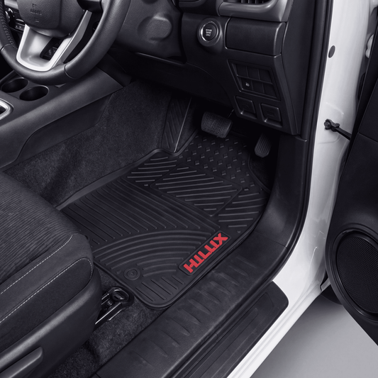 Black rubber car floor mat with a red HILUX logo, shown installed in the driver's footwell of a vehicle interior.