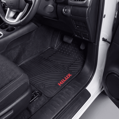 Black rubber car floor mat with a red HILUX logo, shown installed in the driver's footwell of a vehicle interior.
