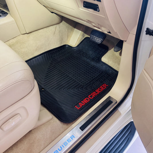 A black rubber car floor mat with red 'LAND CRUISER' text, shown installed in the footwell of a beige vehicle interior.