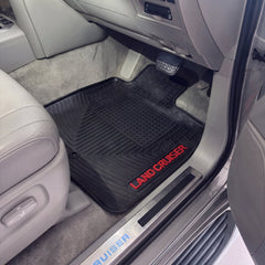 A black rubber car floor mat with red 'LAND CRUISER' lettering, shown installed in the driver's seat of a vehicle with gray leather interior.