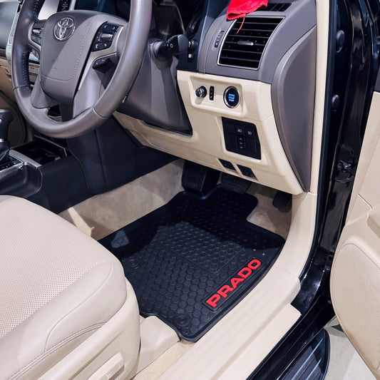 Black rubber car floor mats with red PRADO lettering, shown installed in the front of a beige Toyota vehicle with the door open.