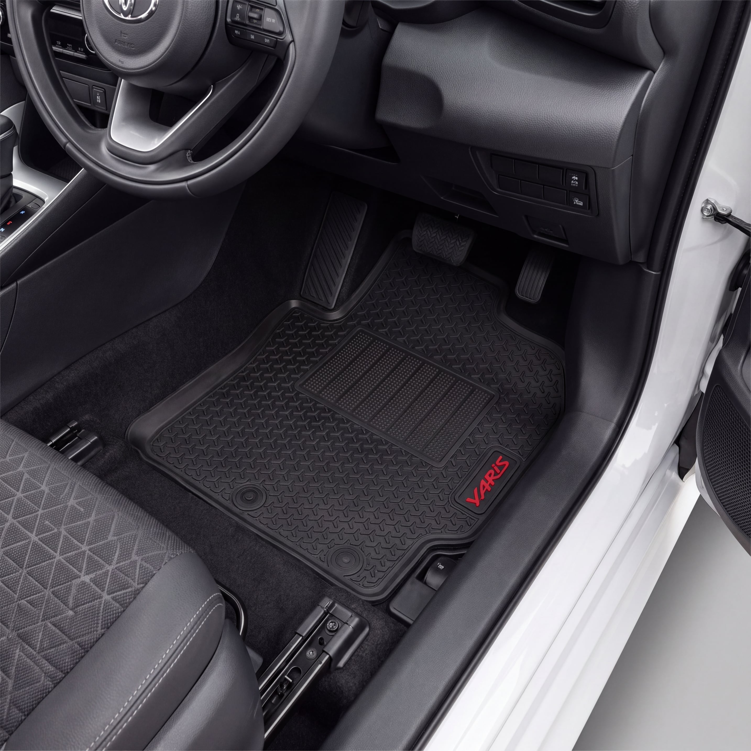 Black car floor mats installed in a vehicle's footwell, with a visible red VARI5 logo on the lower right mat. The photo shows the interior door area, seat, pedals, and dashboard.