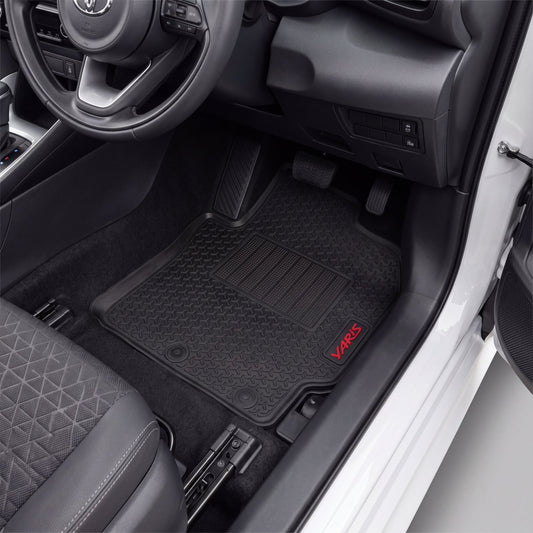 Black car floor mats installed in a vehicle's footwell, with a visible red VARI5 logo on the lower right mat. The photo shows the interior door area, seat, pedals, and dashboard.