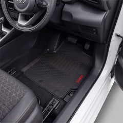 Black car floor mats installed in a vehicle's footwell, with a visible red VARI5 logo on the lower right mat. The photo shows the interior door area, seat, pedals, and dashboard.